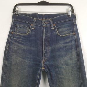 May include: A pair of dark blue denim jeans featuring a classic five-pocket design, a button fly closure, and copper-toned rivets. The fabric displays a faded wash with whiskering patterns around the hips and pockets, suspended on a black hanger.