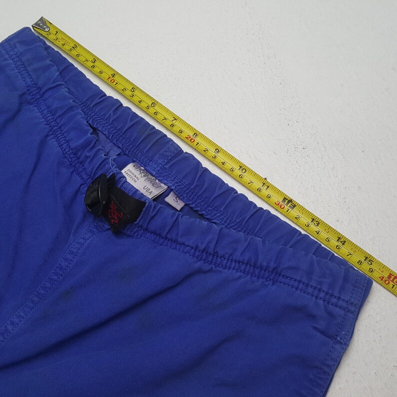 Puede incluir: Primer plano de pantalones azules con cintura el&aacute;stica y hebilla negra. Una cinta m&eacute;trica amarilla muestra la longitud en cent&iacute;metros. Los pantalones tienen una peque&ntilde;a etiqueta que dice "USA".