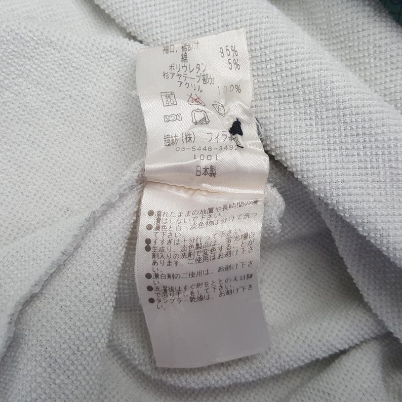 May include: Close-up of a white fabric with a tag attached. The tag has Japanese text and details about the fabric composition: 95% cotton, 5% polyurethane, and 100% acrylic. The tag also includes washing instructions.