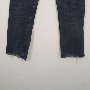 May include: A pair of dark blue denim jeans featuring a straight leg cut and a distressed, frayed raw hem at the bottom of each leg. The fabric displays subtle whiskering and fading patterns along the upper thigh area of the denim material.