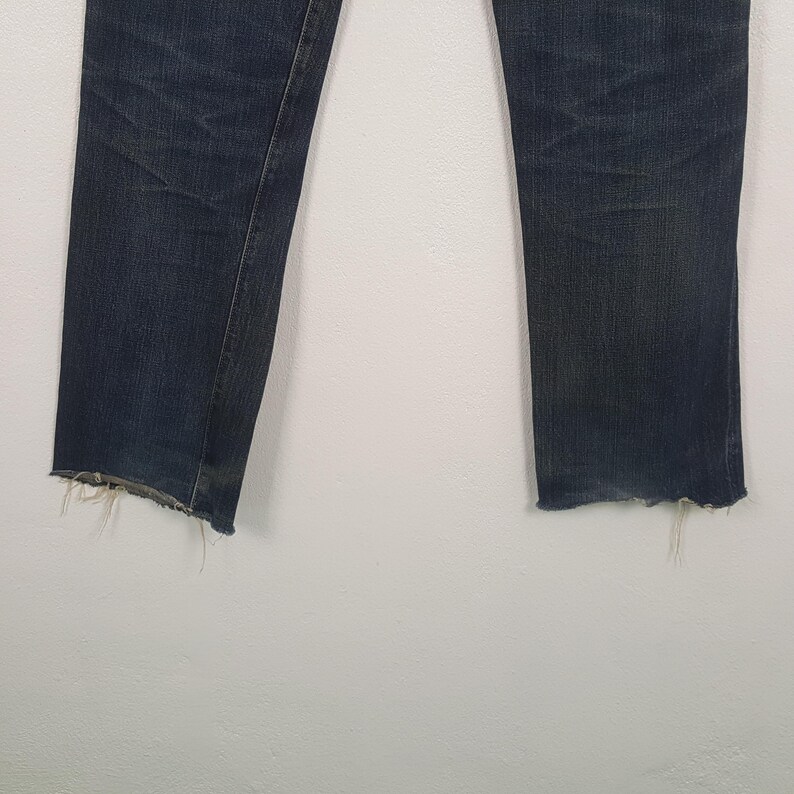 May include: A pair of dark blue denim jeans featuring a straight leg cut and a distressed, frayed raw hem at the bottom of each leg. The fabric displays subtle whiskering and fading patterns along the upper thigh area of the denim material.