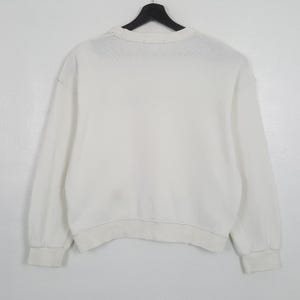 Puede incluir: Una sudadera blanca de cuello redondo y manga larga, colgada de una percha negra. Los pu&ntilde;os y el dobladillo son de canal&eacute;. La tela parece ser un material suave y de peso medio. El fondo es blanco.