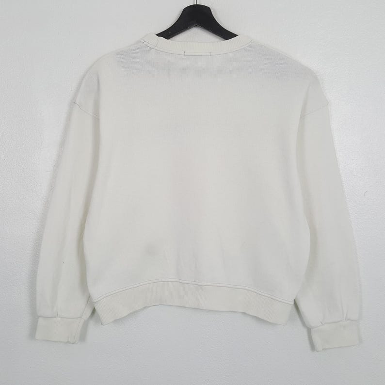 Puede incluir: Una sudadera blanca de cuello redondo y manga larga, colgada de una percha negra. Los pu&ntilde;os y el dobladillo son de canal&eacute;. La tela parece ser un material suave y de peso medio. El fondo es blanco.