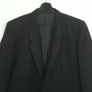 May include: A black blazer hanging on a black hanger against a white background. The blazer has a notched lapel, a chest pocket, and a two-button closure. The interior lining is a dark blue color.