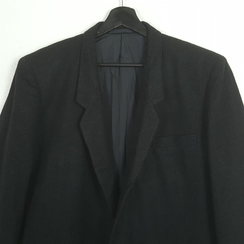 May include: A black blazer hanging on a black hanger against a white background. The blazer has a notched lapel, a chest pocket, and a two-button closure. The interior lining is a dark blue color.