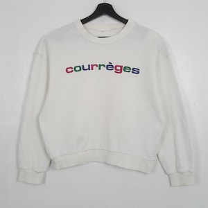 Puede incluir: Sudadera blanca de cuello redondo con la palabra "courr&egrave;ges" bordada en el pecho en rojo, verde, azul y morado. La sudadera de manga larga est&aacute; colgada de una percha negra.