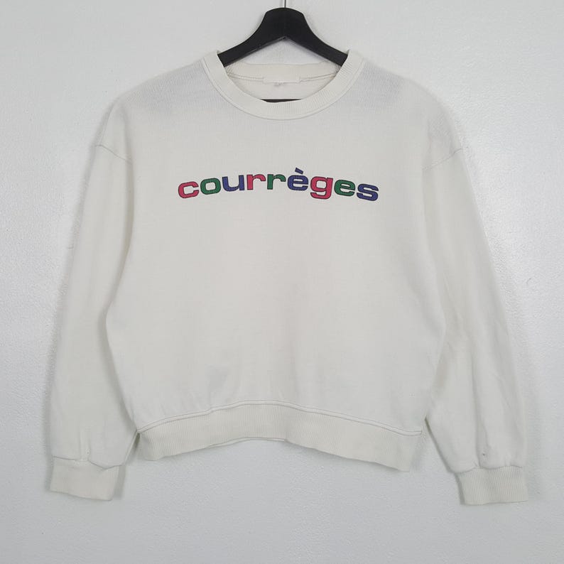 Puede incluir: Sudadera blanca de cuello redondo con la palabra "courr&egrave;ges" bordada en el pecho en rojo, verde, azul y morado. La sudadera de manga larga est&aacute; colgada de una percha negra.