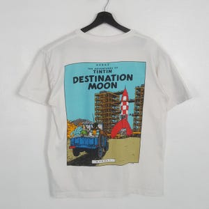 May include: White t-shirt featuring a colorful graphic from "The Adventures of Tintin: Destination Moon." The design includes a rocket, a blue vehicle, and the text "DESTINATION MOON" on a light blue background. The shirt is hanging on a black hanger.