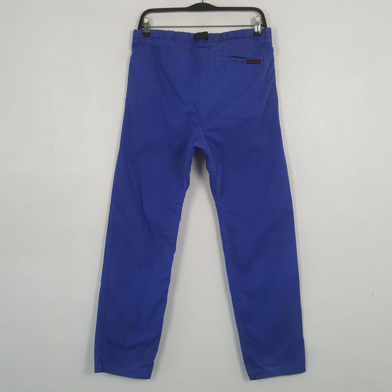 Puede incluir: Un pantal&oacute;n azul colgado de una percha negra. El pantal&oacute;n es de color azul liso y parece estar hecho de una tela duradera. La parte trasera del pantal&oacute;n es visible, mostrando la cintura y un peque&ntilde;o logotipo.