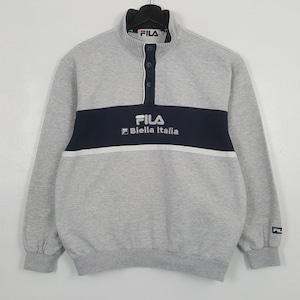 May include: A grey Fila sweatshirt with a navy blue stripe across the chest, displaying the brand's logo and "Biella Italia" in white. The sweatshirt has a button-up collar and long sleeves.