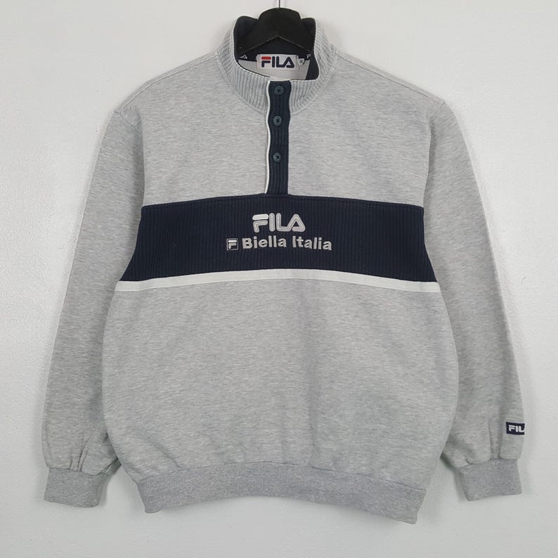 May include: A grey Fila sweatshirt with a navy blue stripe across the chest, displaying the brand's logo and "Biella Italia" in white. The sweatshirt has a button-up collar and long sleeves.