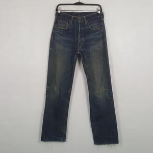 May include: A pair of dark blue denim jeans featuring a high-waisted fit, a button fly closure, and a straight-leg silhouette. The fabric displays a faded wash effect on the thighs and knees, with raw, frayed edges at the bottom hems.
