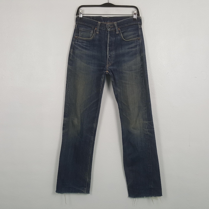 May include: A pair of dark blue denim jeans featuring a high-waisted fit, a button fly closure, and a straight-leg silhouette. The fabric displays a faded wash effect on the thighs and knees, with raw, frayed edges at the bottom hems.