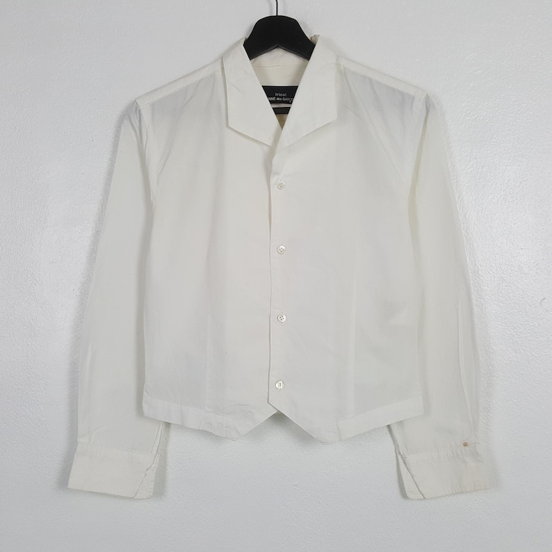 May include: A white, long-sleeved shirt with a unique collar and button-down front. The shirt has a tailored fit and is made of a lightweight fabric. The cuffs are buttoned. The shirt is hanging on a black hanger.