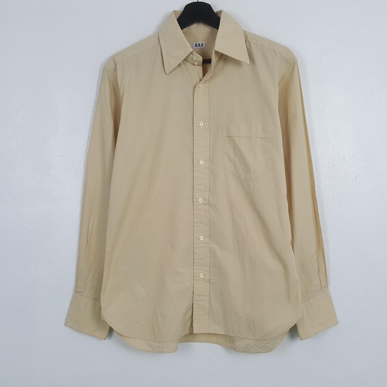 Pode incluir: Uma camisa de manga comprida com bot&otilde;es numa cor bege claro s&oacute;lida. A camisa apresenta um colarinho pontiagudo, um bolso &uacute;nico no peito no lado esquerdo e uma carcela frontal com bot&otilde;es bege a condizer. A etiqueta da marca A.A.R &eacute; vis&iacute;vel.