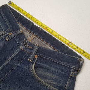 May include: Dark blue denim jeans featuring a metal button closure, front pockets, and visible stitching. A yellow measuring tape is placed along the waistband, indicating a measurement of approximately 14.75 inches across the top edge.
