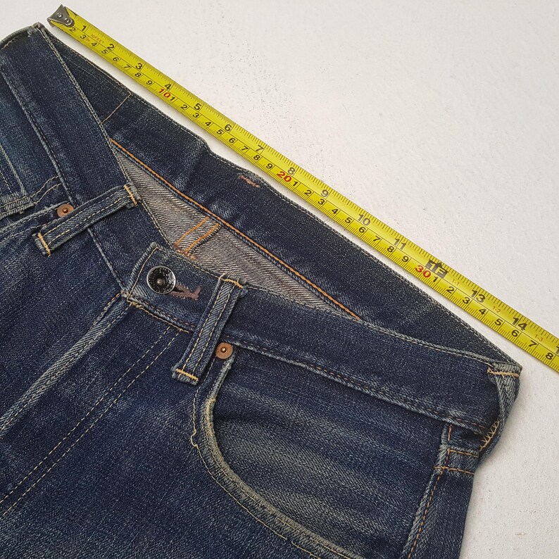 May include: Dark blue denim jeans featuring a metal button closure, front pockets, and visible stitching. A yellow measuring tape is placed along the waistband, indicating a measurement of approximately 14.75 inches across the top edge.