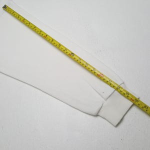 Puede incluir: Manga de camisa blanca de manga larga con pu&ntilde;o acanalado. Una cinta m&eacute;trica amarilla est&aacute; colocada sobre la manga, mostrando medidas en cent&iacute;metros. La manga est&aacute; hecha de un material suave y opaco.