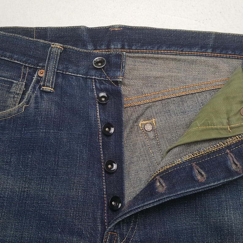 May include: Dark blue denim jeans featuring a button-fly closure with five black buttons. The interior pocket lining is olive green fabric. The jeans have visible orange contrast stitching along the seams and a small metal rivet on the pocket.