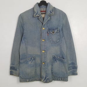 May include: Light blue denim jacket with a classic collar, four gold-colored buttons, and two large front pockets. Features a small chest pocket with a red tag. The jacket is hanging on a black hanger.