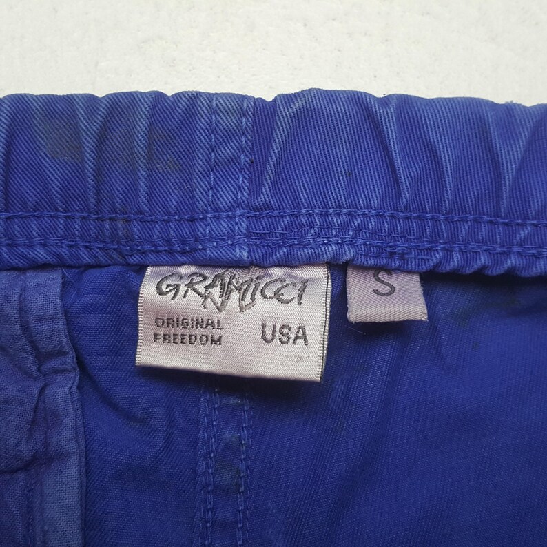 Puede incluir: Primer plano de unos pantalones cortos Gramicci azules con una etiqueta blanca. La etiqueta dice "GRAMICCI ORIGINAL FREEDOM USA" en texto negro. Tambi&eacute;n es visible una peque&ntilde;a etiqueta blanca con la letra "S". Los pantalones cortos est&aacute;n hechos de tela duradera.