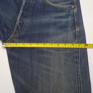 May include: Dark blue denim jeans featuring a faded wash and copper-toned rivets. A yellow measuring tape is positioned across the upper thigh area, showing a width measurement of approximately 12 inches from the side seam to the crotch seam.