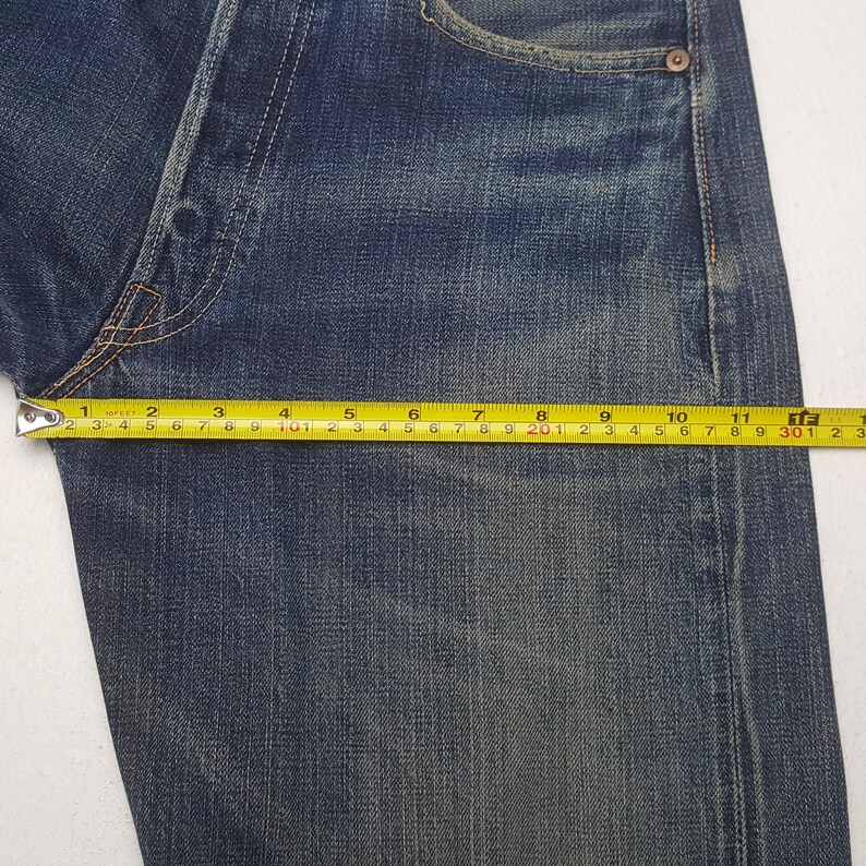 May include: Dark blue denim jeans featuring a faded wash and copper-toned rivets. A yellow measuring tape is positioned across the upper thigh area, showing a width measurement of approximately 12 inches from the side seam to the crotch seam.