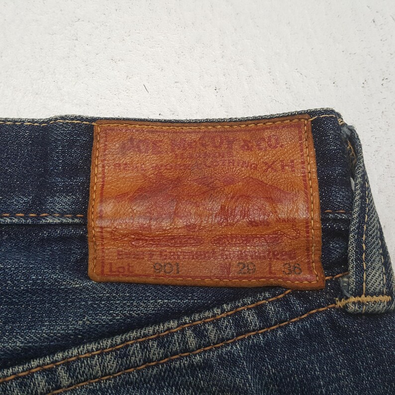 May include: Dark blue denim jeans featuring a rectangular tan leather patch stitched onto the waistband. The patch displays embossed text reading The Real McCoy's Co., Lot 901, W 29, and L 36, along with a graphic illustration of two mountains.