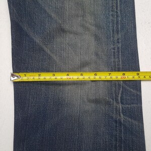 May include: A pair of dark blue denim jeans featuring a faded wash and visible stitching. A yellow measuring tape is placed horizontally across the fabric, indicating a width measurement of approximately 9.5 inches across the denim material.