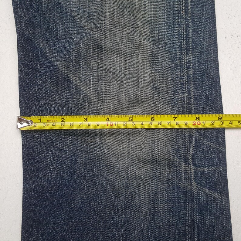 May include: A pair of dark blue denim jeans featuring a faded wash and visible stitching. A yellow measuring tape is placed horizontally across the fabric, indicating a width measurement of approximately 9.5 inches across the denim material.