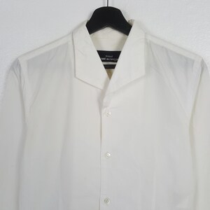 May include: A white button-down shirt with a wide collar, hanging on a black hanger. The shirt has a minimalist design with a few visible buttons down the front. The label reads "tricot COMME des GAR&Ccedil;ON".