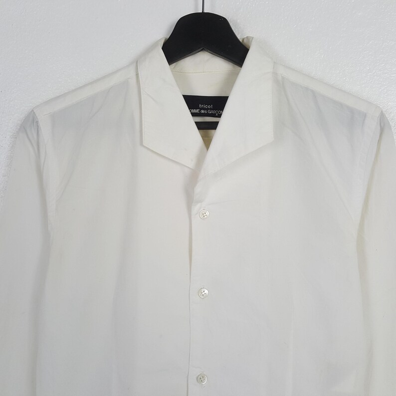 May include: A white button-down shirt with a wide collar, hanging on a black hanger. The shirt has a minimalist design with a few visible buttons down the front. The label reads "tricot COMME des GAR&Ccedil;ON".