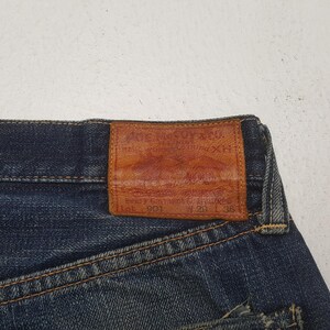 May include: Dark blue denim jeans featuring a rectangular tan leather patch on the back waistband. The patch is embossed with the text The Real McCoy's & Co. Real Work Clothing XH, Lot 901, W 29 L 36. The jeans have visible orange stitching.