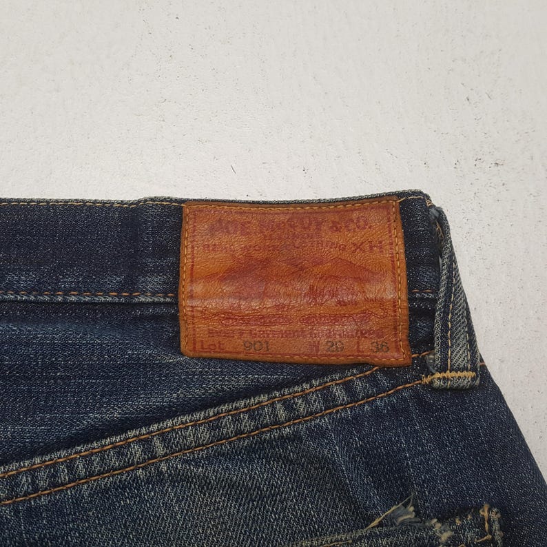 May include: Dark blue denim jeans featuring a rectangular tan leather patch on the back waistband. The patch is embossed with the text The Real McCoy's & Co. Real Work Clothing XH, Lot 901, W 29 L 36. The jeans have visible orange stitching.