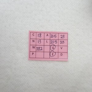 Puede incluir: Una peque&ntilde;a etiqueta rectangular rosa con texto y n&uacute;meros negros escritos a mano. La etiqueta tiene una cuadr&iacute;cula de casillas con letras y n&uacute;meros, incluyendo "C 18", "A 21-5" y "W 382". La etiqueta est&aacute; sobre una superficie blanca texturizada.