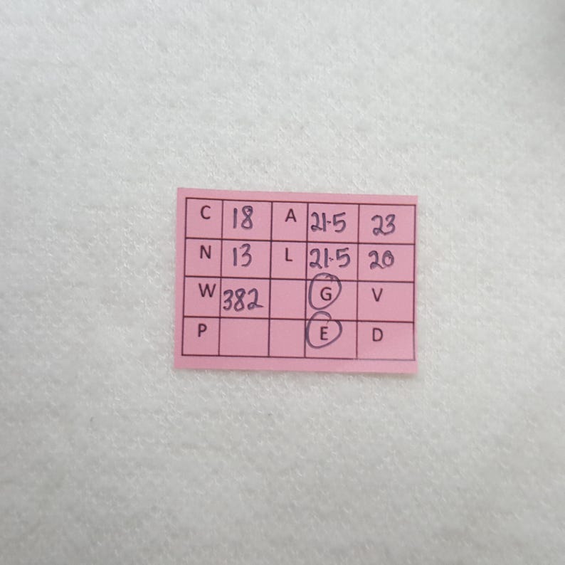 Puede incluir: Una peque&ntilde;a etiqueta rectangular rosa con texto y n&uacute;meros negros escritos a mano. La etiqueta tiene una cuadr&iacute;cula de casillas con letras y n&uacute;meros, incluyendo "C 18", "A 21-5" y "W 382". La etiqueta est&aacute; sobre una superficie blanca texturizada.