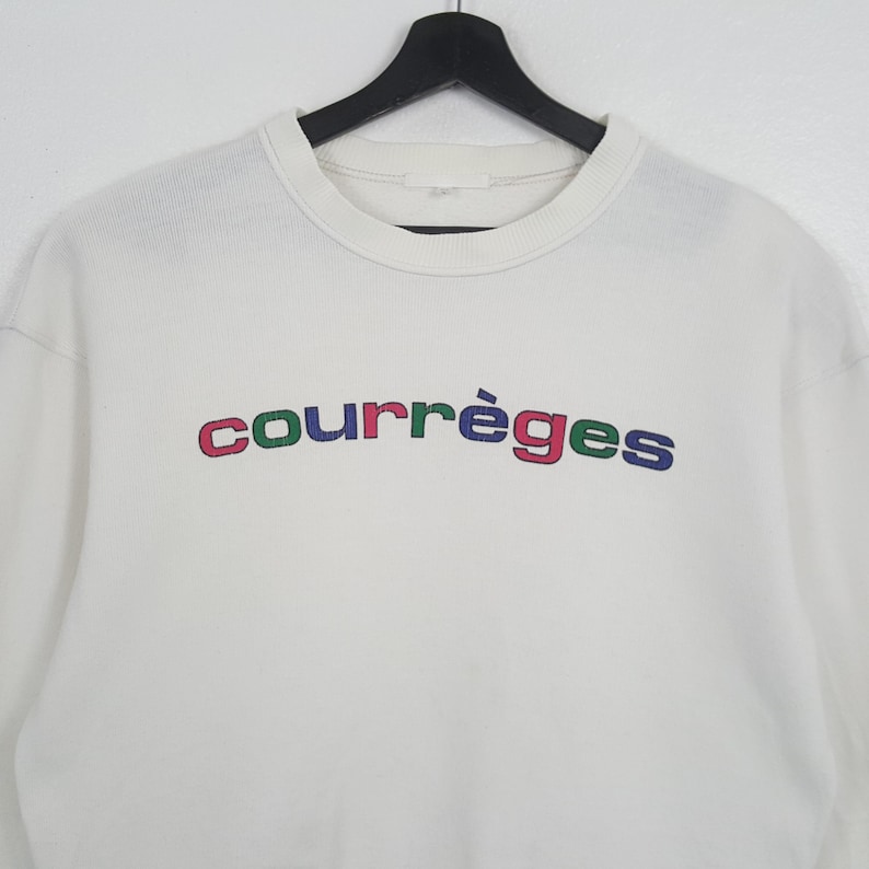 Puede incluir: Una sudadera blanca con la palabra "courr&egrave;ges" en letras multicolores en el pecho. El texto es rojo, verde, azul y morado. La sudadera tiene cuello redondo y mangas largas.