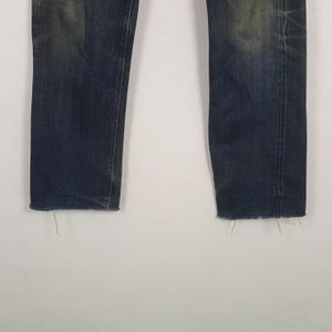 May include: A pair of dark blue denim jeans featuring a faded wash on the thighs and knees. The legs are straight-cut with raw, frayed edges at the bottom hems, showing loose threads and a worn-in appearance throughout the fabric surface.