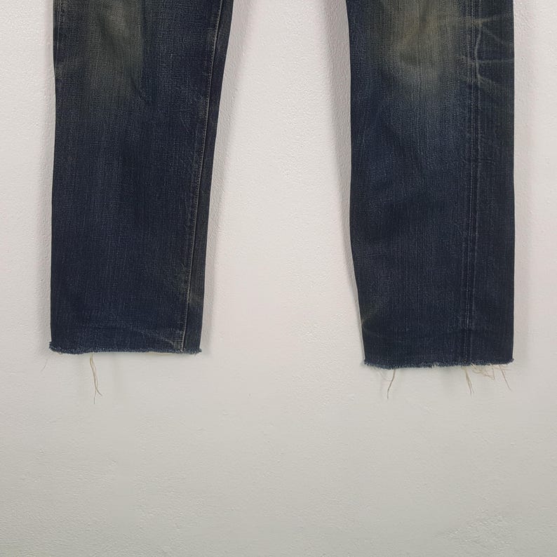 May include: A pair of dark blue denim jeans featuring a faded wash on the thighs and knees. The legs are straight-cut with raw, frayed edges at the bottom hems, showing loose threads and a worn-in appearance throughout the fabric surface.