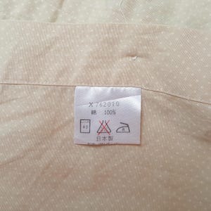 Pode incluir: Um tecido bege com padr&atilde;o de pontos brancos e uma etiqueta de cuidados branca. A etiqueta exibe o texto X 762010, 100% algod&atilde;o e caracteres japoneses para fabricado no Jap&atilde;o, junto com s&iacute;mbolos de lavagem, branqueamento e engomagem.