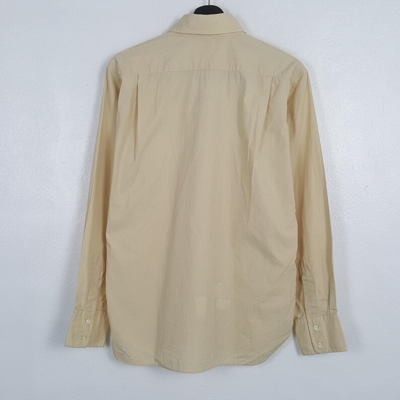 Pode incluir: Uma camisa de manga comprida com bot&otilde;es em um tom bege claro s&oacute;lido. A camisa apresenta uma gola padr&atilde;o, uma pala traseira com uma prega central e punhos com dois bot&otilde;es vis&iacute;veis em cada manga. O tecido parece liso e leve.