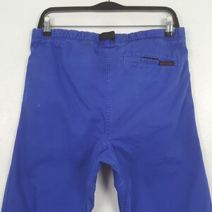 Puede incluir: Pantalones azul brillante con cintura el&aacute;stica y hebilla negra. La parte trasera presenta un bolsillo con un peque&ntilde;o logotipo rojo y blanco. Los pantalones cuelgan de una percha negra sobre un fondo blanco.