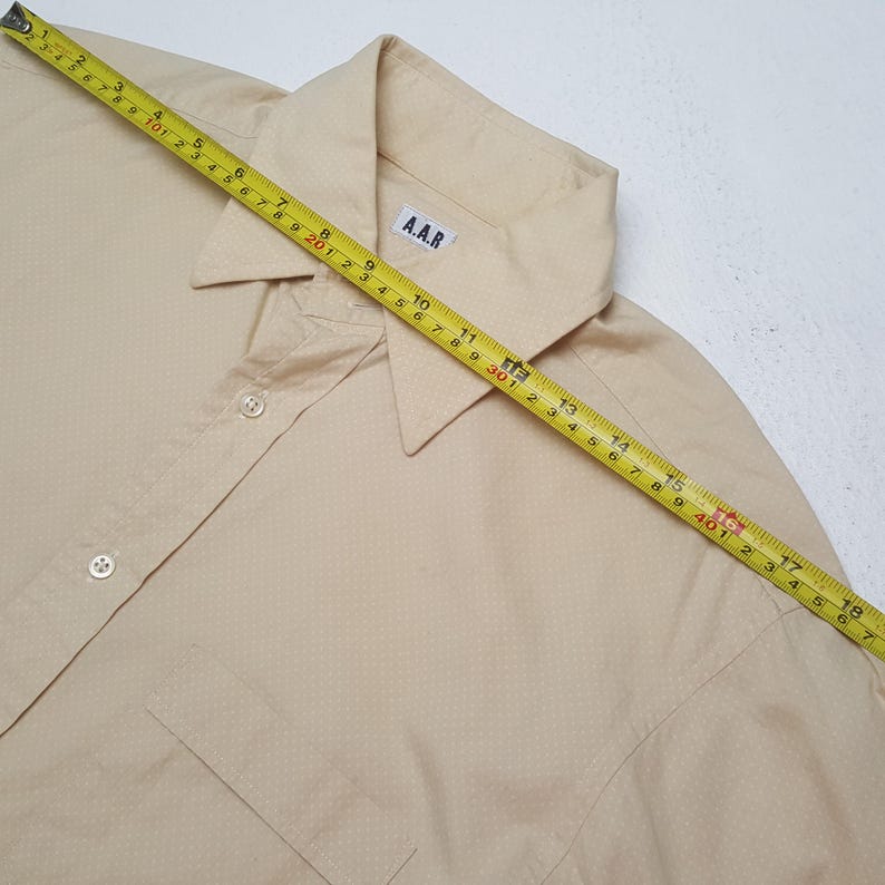 Pode incluir: Uma camisa de bot&otilde;es bege claro com um padr&atilde;o subtil de pontos brancos, colarinho pontiagudo e uma etiqueta A.A.R. Uma fita m&eacute;trica amarela est&aacute; colocada diagonalmente sobre a camisa, mostrando um comprimento de cerca de 46 cm.