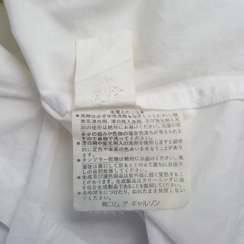 May include: A white fabric tag with Japanese text, likely a care label. The tag is attached to a white garment, with visible text detailing washing instructions. The tag is rectangular with rounded corners.