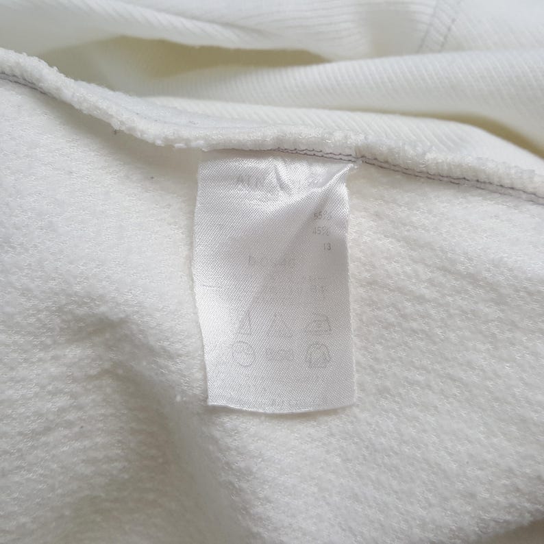 Puede incluir: Primer plano de una tela blanca con una etiqueta blanca adjunta. La etiqueta tiene texto y s&iacute;mbolos negros. La tela parece ser un material suave y texturizado, posiblemente una sudadera o una prenda similar.