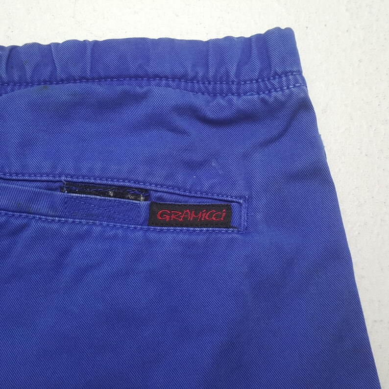 Puede incluir: Primer plano de pantalones Gramicci azules. Los pantalones tienen una cintura el&aacute;stica y un bolsillo trasero con el logotipo de Gramicci en rojo. La tela parece ser una mezcla de algod&oacute;n duradera.
