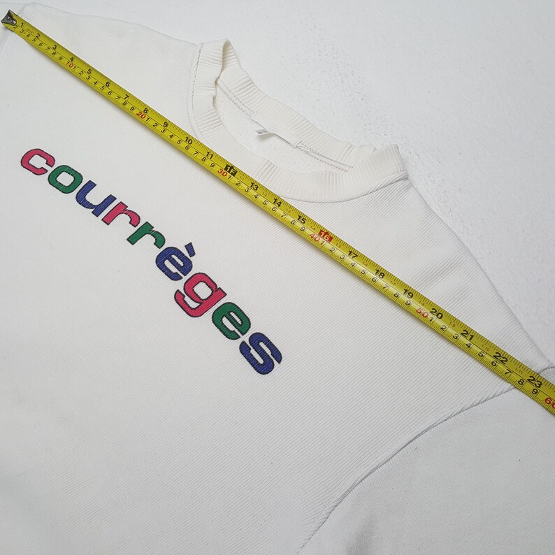 Puede incluir: Sudadera blanca de cuello redondo con la palabra "courr&egrave;ges" impresa en letras coloridas. El texto est&aacute; en un dise&ntilde;o diagonal. Una cinta m&eacute;trica amarilla est&aacute; colocada sobre la sudadera.