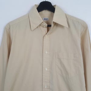 Pode incluir: Uma camisa de manga comprida bege claro com bot&otilde;es e um padr&atilde;o subtil de micro-pontos. A camisa apresenta um colarinho pontiagudo, um bolso &uacute;nico no peito do lado esquerdo e uma etiqueta branca no interior da gola com o texto A.A.R.