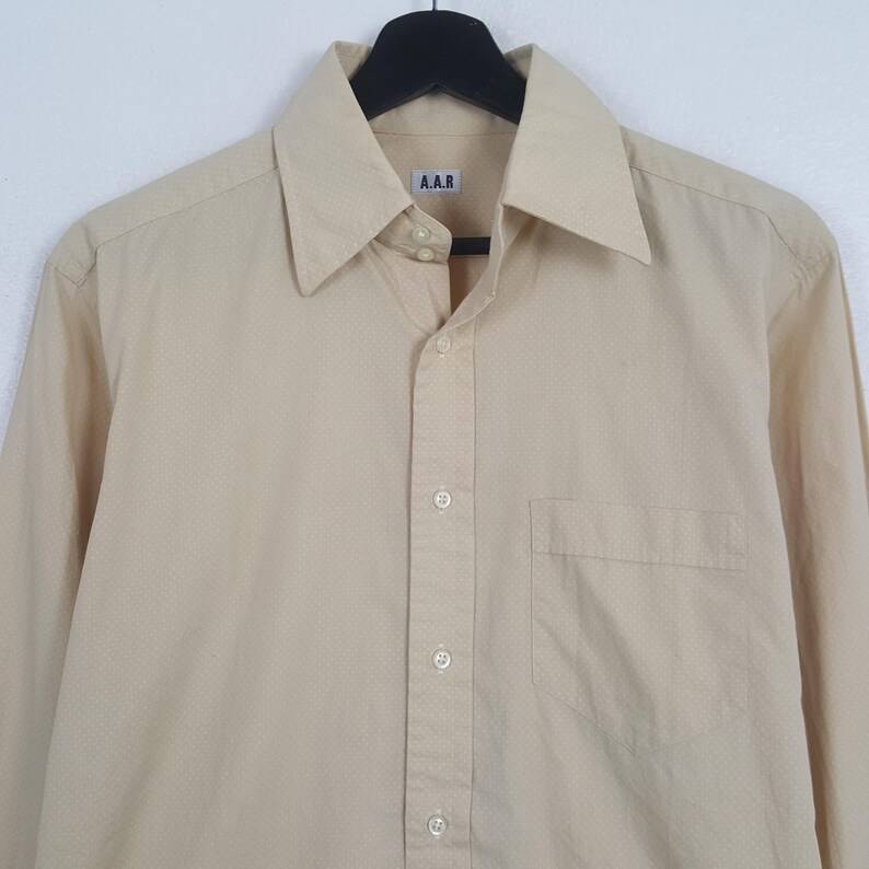 Pode incluir: Uma camisa de manga comprida bege claro com bot&otilde;es e um padr&atilde;o subtil de micro-pontos. A camisa apresenta um colarinho pontiagudo, um bolso &uacute;nico no peito do lado esquerdo e uma etiqueta branca no interior da gola com o texto A.A.R.