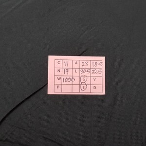May include: A close-up of a dark gray fabric with a pink tag attached. The tag has handwritten text and numbers, including "C 11", "A 23", "N 19", "L 30.5", and "W 1000". The fabric has a subtle texture.
